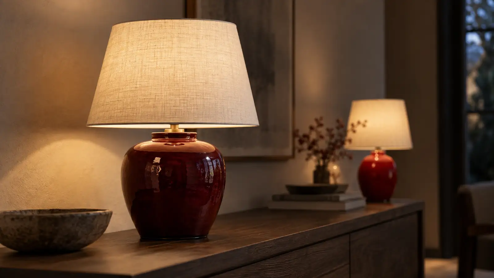 Bordslampa med vinröd glaserad fot och ljus linneskärm på ett mörkt ekbord, med en klarröd lampa suddig i bakgrunden